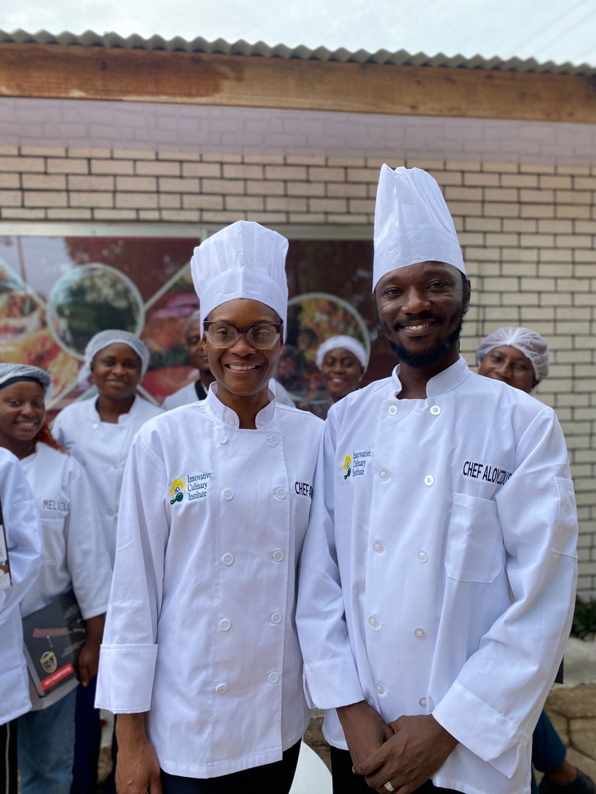 Chef Adanne and Chef Aloysious in chef hats with participants behind